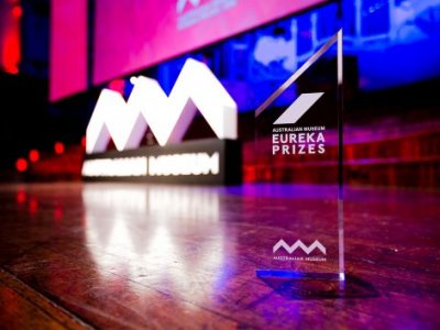 A photo of a stage, lit up with pink and blue lights, and a glass trophy in the foreground saying Eureka Prizes.